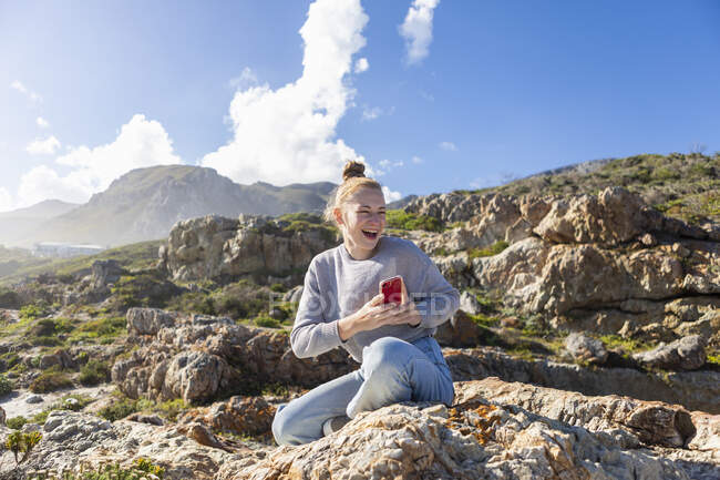 Девочка-подросток фотографирует со своего смартфона, сидит на скалах на пляже — стоковое фото