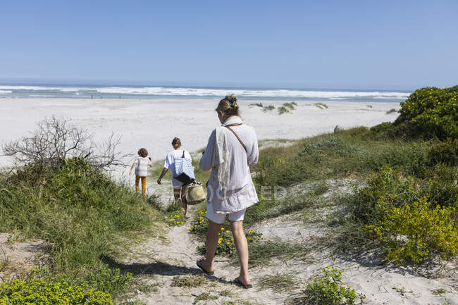 Una familia caminando a través de las dunas de arena hacia el océano con cestas y bolsas. - foto de stock