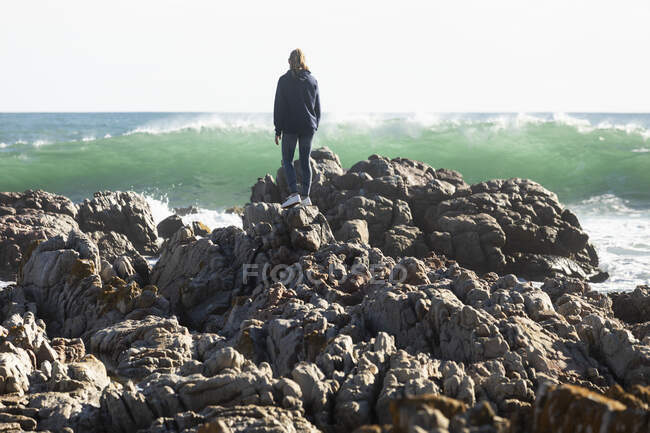 Adolescente escalando en las rocas dentadas en una playa, grandes olas rompiendo en la orilla - foto de stock
