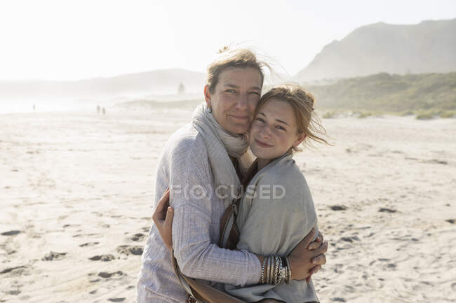 Mulher adulta e sua filha adolescente abraçando, de pé em uma praia varrida pelo vento — Fotografia de Stock