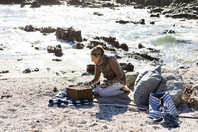 Mujer adulta en una dorada y bufanda con una cesta de picnic y alfombras en una playa de arena junto al agua. - foto de stock