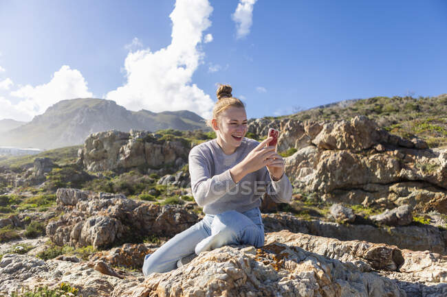 Adolescente prenant des photos avec son téléphone intelligent, assise sur les rochers sur une plage — Photo de stock