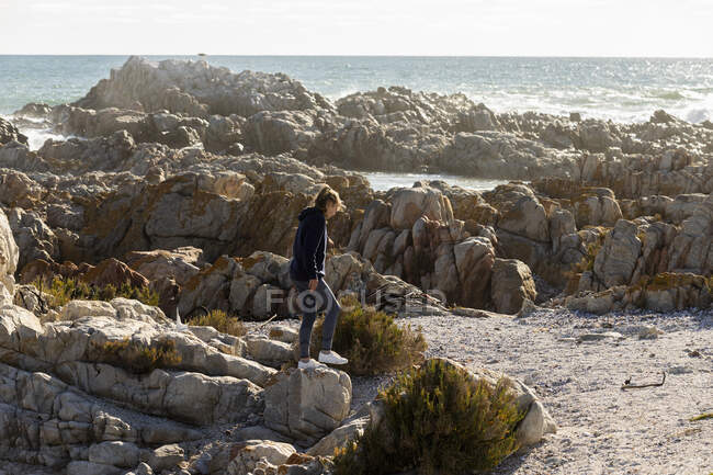 Девочка-подросток карабкается по неровным скалам на пляже, большие волны ломаются на берегу — стоковое фото