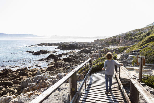 A boy walking along a seashore boardwalk above the rocks. — Stock Photo