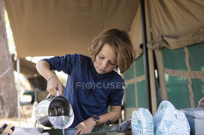 Un ragazzo che versa acqua in un bacino in un campo safari, lavandosi. — Foto stock