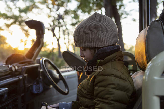 Um rapaz de chapéu e casaco num jipe ao nascer do sol numa viagem de safári. — Fotografia de Stock