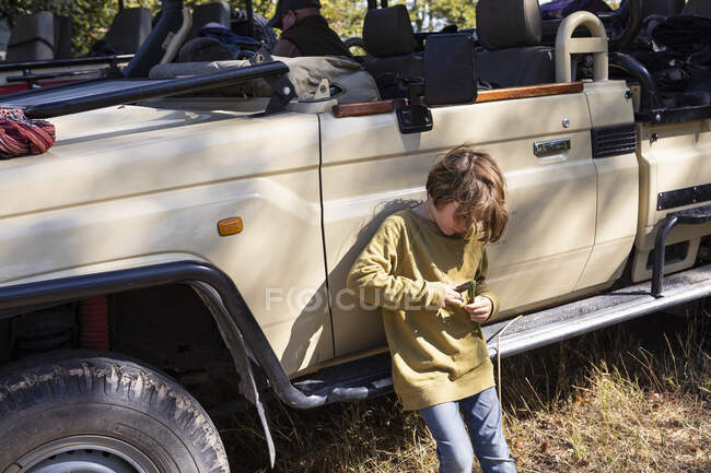 Eight year old boy leaning against a jeep, playing with a toy. — Stock Photo