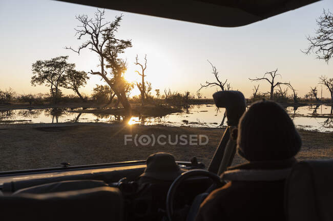 Persona seduta in una jeep a guardare il sole sorgere sull'acqua. — Foto stock