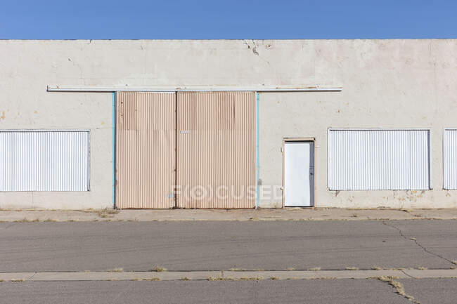 Bâtiment d'entrepôt fermé avec volets métalliques sur les portes et les fenêtres, et mauvaises herbes poussant à travers l'aire de trafic. — Photo de stock