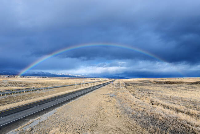 An interstate road reaching to the horizon through a flat landscape, rainbow above the road. — Fotografia de Stock