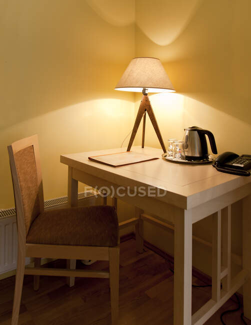 Номер с небольшим письменным столом и стулом с зажженной настольной лампой, подносом и чайником и телефоном. — стоковое фото