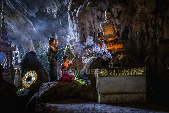 A cave temple with Buddha statues, lit candles and two girls kneeling and praying. — Stock Photo