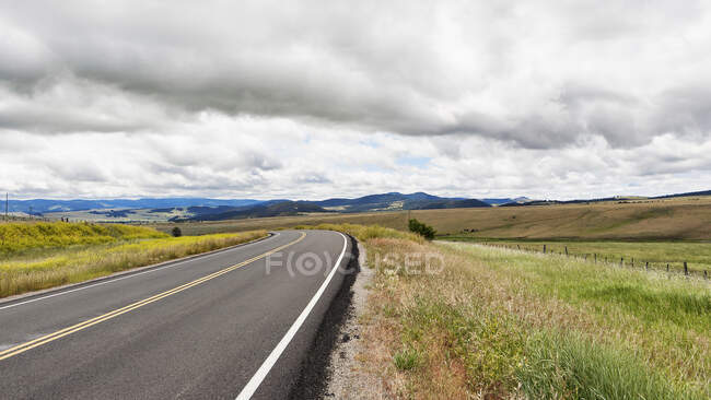 A blacktop road through a flat landscape with undulating hills, farmland and rolling hills beyond. — Stock Photo