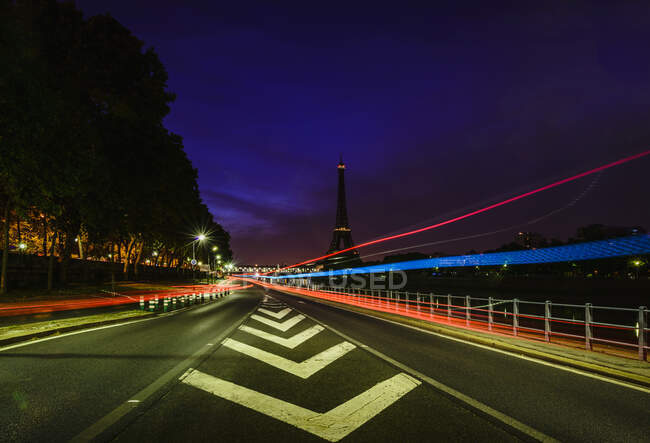 A view along a city road at night, tall buildings and light trails, the Eiffel tower in the distance. — Stock Photo