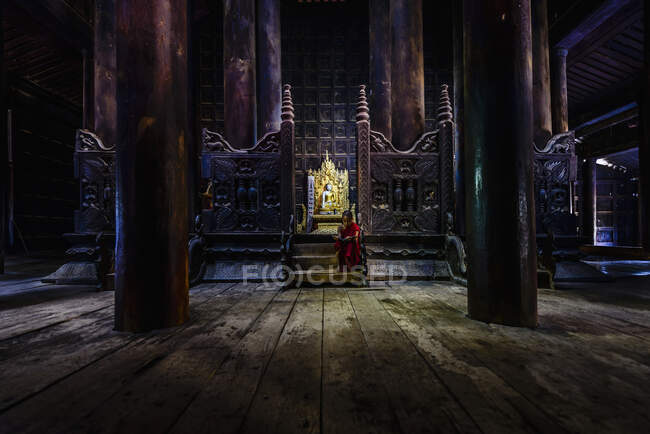 Un templo budista con pilares de madera y pantallas talladas, un tronco dorado y un joven monje sentado en las escaleras. - foto de stock