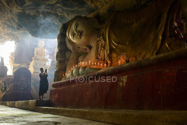 Um templo de caverna, uma grande estátua de Buda reclinada, velas acesas e um monge orando. — Fotografia de Stock