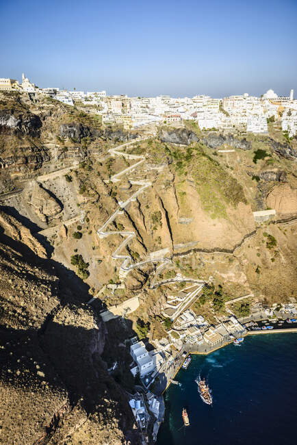 Elevated view of a steep cliff, hilltop town and a harbour on the water with a zigzag path up the cliffside on a Greek island. — Stock Photo
