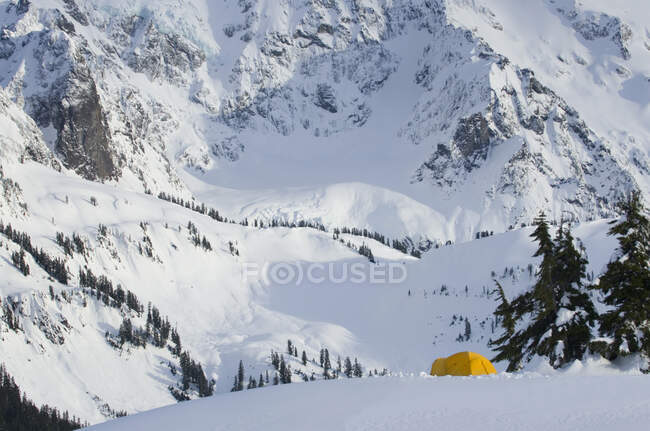 A small yellow tent pitched in deep snow on a slope, view of the steep slopes of the mountains. — Stock Photo