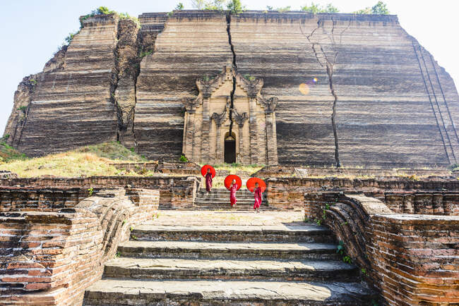 Three monks with orange umbrellas outside a large rock temple, the entrance carved into the rock face at Saigang. — Stock Photo