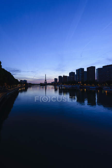 A view along the water of the River Seine at night, tall buildings on the river bank, the Eiffel tower in the distance. — Stock Photo
