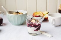 Glass of yoghurt with jam on kitchen table with fruit and granola — Stock Photo