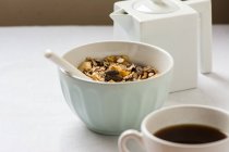 Tigela com granola e colher por xícara de café na mesa branca — Fotografia de Stock