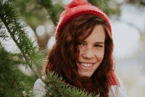 Portrait d'une fille aux taches de rousseur souriante en bonnet tricoté rouge parmi les sapins — Photo de stock