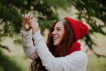 Portrait of laughing ginger girl in knitted red hat decorating fir tree outside — Stock Photo