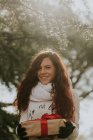 Портрет улыбающейся рыжей девушки, держащей подарок, смотрящей в камеру в зимнем лесу — стоковое фото