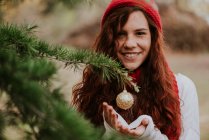 Portrait of smiling redhead girl holding hands under bauble hanging on pine tree. — Stock Photo