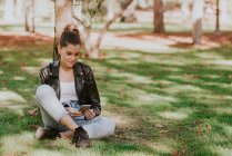 Портрет улыбающейся девушки, сидящей на траве и просматривающей смартфон — стоковое фото