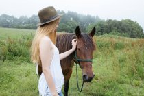 Chica con sombrero acariciando caballo marrón - foto de stock