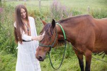 Брюнетка носіння біле плаття стоїть біля коричневий кінь на родовищі — стокове фото