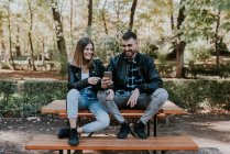 Sonriente pareja sentada en la mesa del parque y el teléfono inteligente de navegación - foto de stock