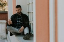 Молодой человек сидит на подоконнике и просматривает смартфон — стоковое фото