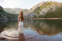 Visão traseira da menina loira vestindo vestido branco em pé no lago da montanha — Fotografia de Stock