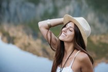 Portrait of brunette girl wearing hat posing with closed eyes over mountain lake — Stock Photo