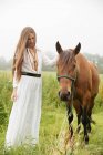 Fille en robe blanche debout près du cheval brun au champ — Photo de stock