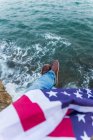 Homem sentado ao lado do mar com uma bandeira dos EUA nas pernas — Fotografia de Stock