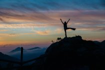 Silhouette de femelle avec les bras et la jambe vers le haut posant sur le sommet de la montagne sur le ciel crépusculaire sur le fond — Photo de stock