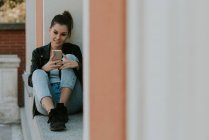 Молодая девушка сидит на подоконнике и печатает на смартфоне — стоковое фото
