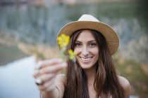 Portrait of brunette woman in hat showing yellow flower at camera against of lake on background — Stock Photo