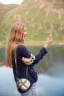Вид збоку білява дівчина з колбою, що звисає на плечі, дивлячись на компас в руці над озером на фоні — стокове фото