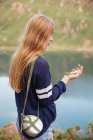 Side view of young girl looking at compass with flask on shoulder at mountain lake — Stock Photo