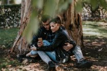 Abrazar a la pareja sentada en el suelo por el árbol y el teléfono inteligente de navegación - foto de stock