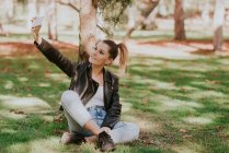 Jovencita alegre sentada junto al árbol en el césped del parque y tomando selfie - foto de stock