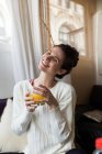 Portrait of smiling girl posing on coach with eyes closed and holding glass of orange juice — Stock Photo