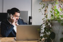 Homme d'affaires confiant assis à son ordinateur portable et parlant au téléphone. Plan intérieur horizontal. — Photo de stock
