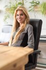Cheerful blond girl sitting at workplace and looking at camera — Stock Photo