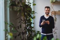 Portrait of smiling man with smartphone standing near window in office and looking at camera — Stock Photo